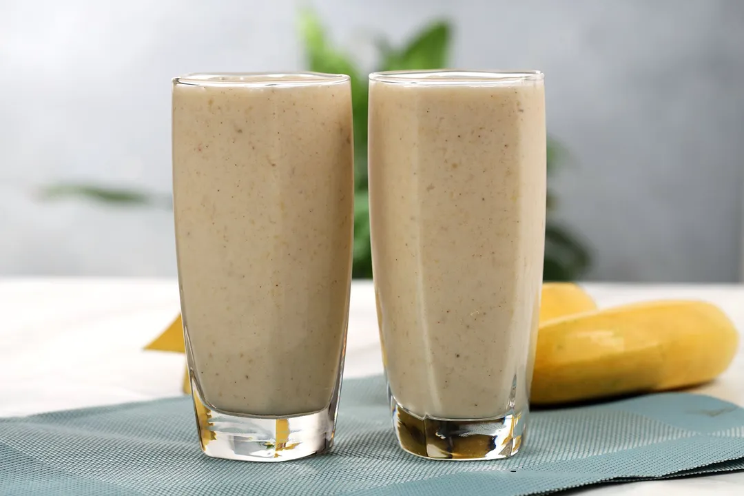 Two glasses of Peanut Butter Banana Smoothie  placed on a blue cloth with some unpeeled yellow bananas in the back.