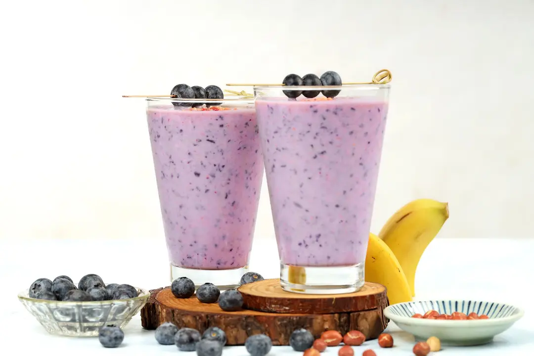 Two glasses of blueberry banana smoothie topped with blueberries and surrounded by extra berries and peanuts