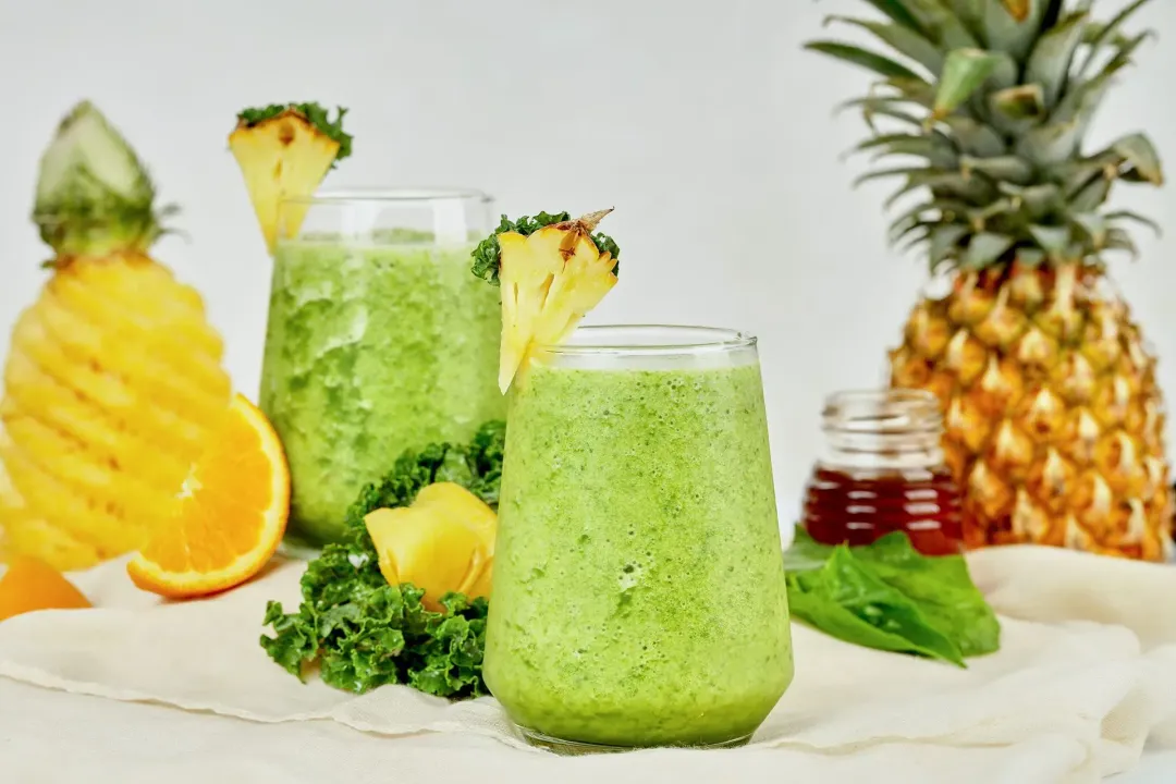 a shot of two pineapple green smoothie glasses taken with a whole pineapple, a jar of honey, some kale leaves, and a peeled pineapple 