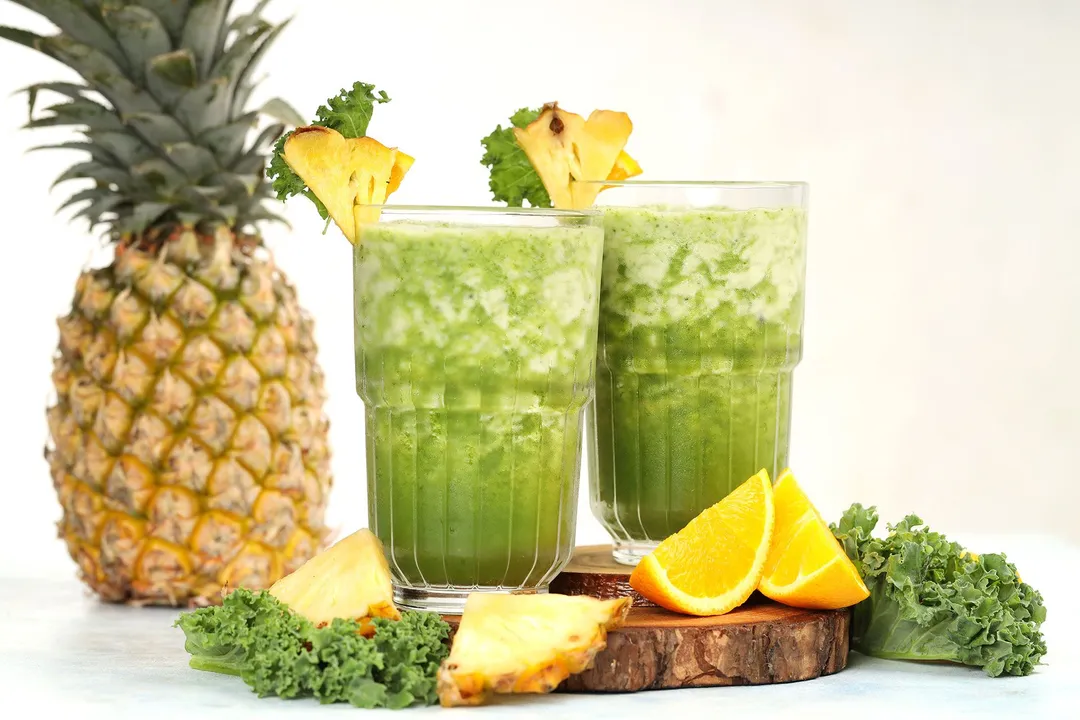 Two glasses of Pineapple Kale Smoothie placed on a wooden board near a pineapple, orange wedges, kale, and pineapple slices.