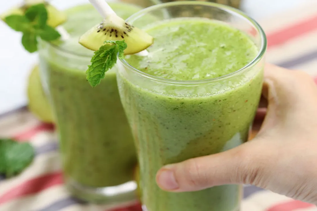 A hand picking up a glass of Pineapple Kiwi Smoothie that's placed next to an identical glass.