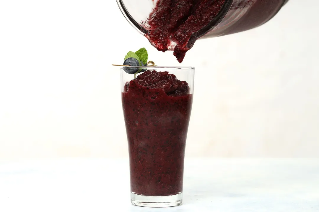 A blender pouring blueberry smoothie into a tall glass.