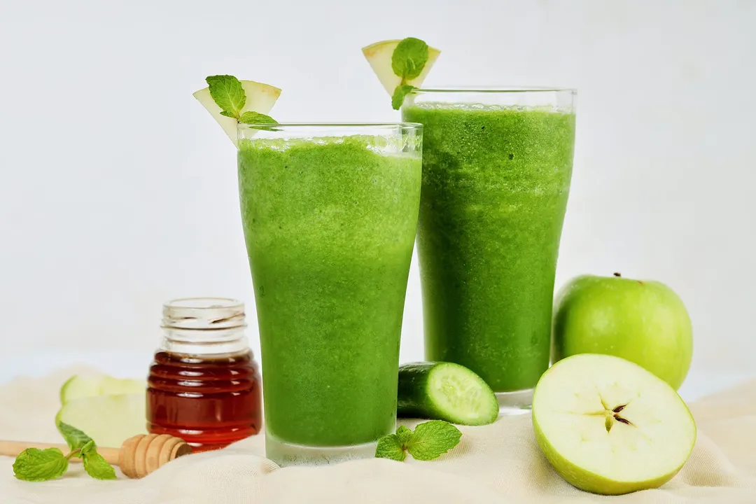 Two tall glasses of sour apple smoothie laid near green apples, cucumbers, and a small jar of honey.