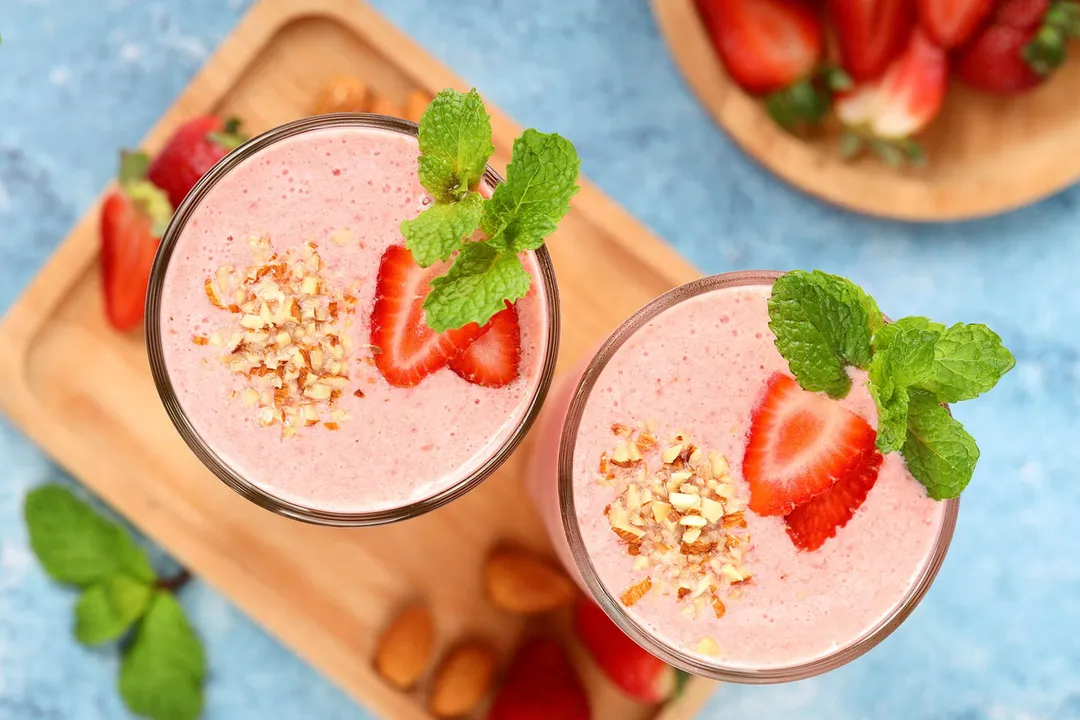 A high-angle shot of two strawberry smoothie glasses laid on a wooden board and near a bowl of strawberry and mint leaves.