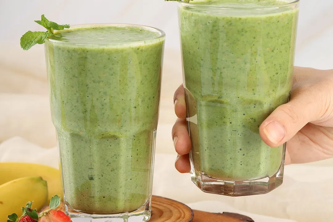 A hand picking up a glass of strawberry spinach banana smoothie placed next to an identical glass.