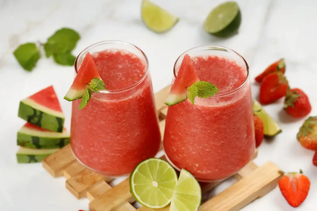 Two glasses of watermelon mojito smoothies placed side by side on a wooden tray surrounded by strawberries, watermelon slices, lime, and mint leaves