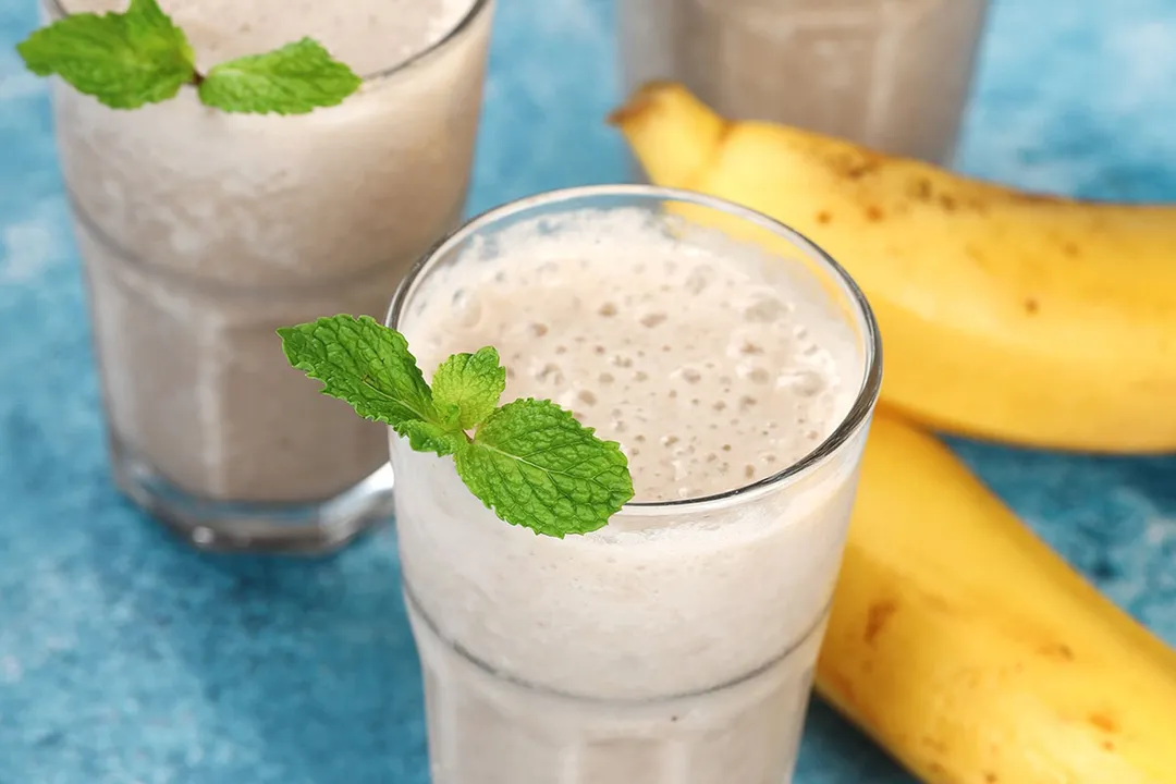 Three glasses of banana smoothie laid near two unpeeled yellow bananas.