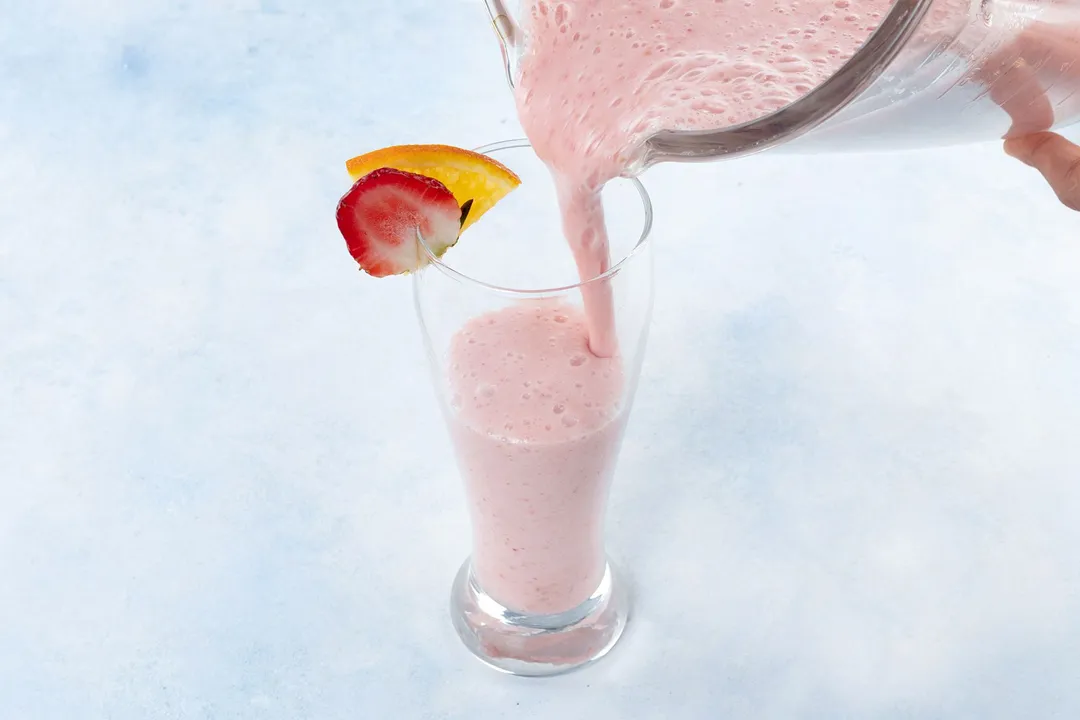 a pitcher pouring pink smoothie into a glass garnished with sliced orange and strawberry