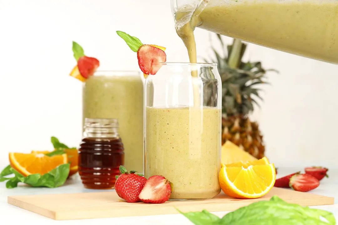 a blender pitcher of smoothie pouring into glasses, surrounded by honey jar, orange sliced, strawberries, pineapple