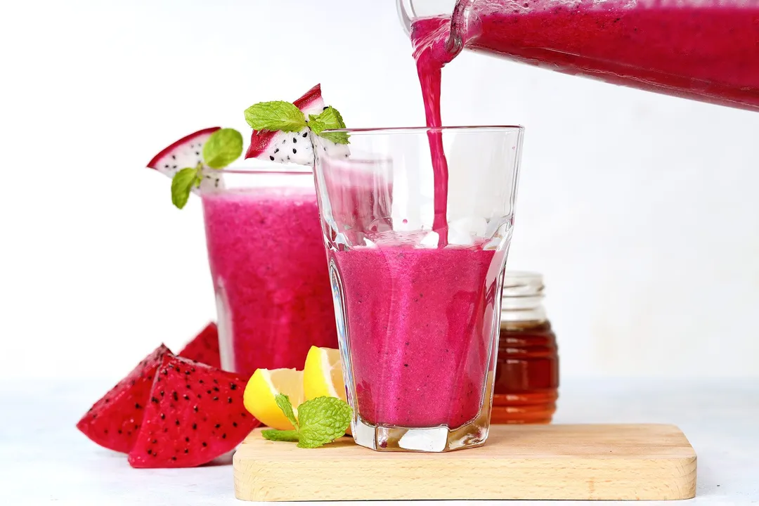 a pitcher pouring pink smoothie into two glasses, decorated with dragon fruit sliced, lemon sliced, honey jar and mint leaves