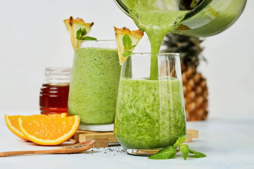 a pitcher pouring green smoothie into two glasses, decorated with orange sliced, pineapple, honey jar
