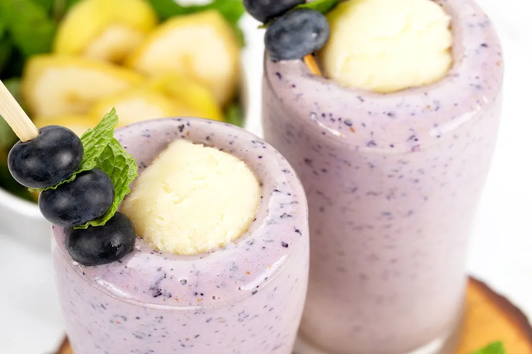 Two glasses of blueberry banana smoothie placed on a wooden board and garnished with blueberry skewers and ice cream scoops.