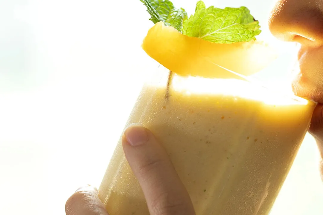 A person sipping on a glass of peach smoothie garnished with a slice of peach and mint leaves.