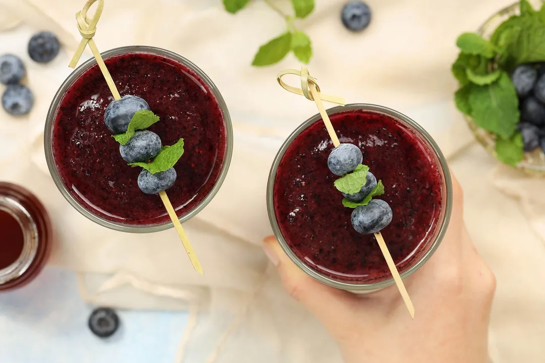 A high angle shot of two glasses of blueberry smoothie, one of which is about to be picked up by a hand.