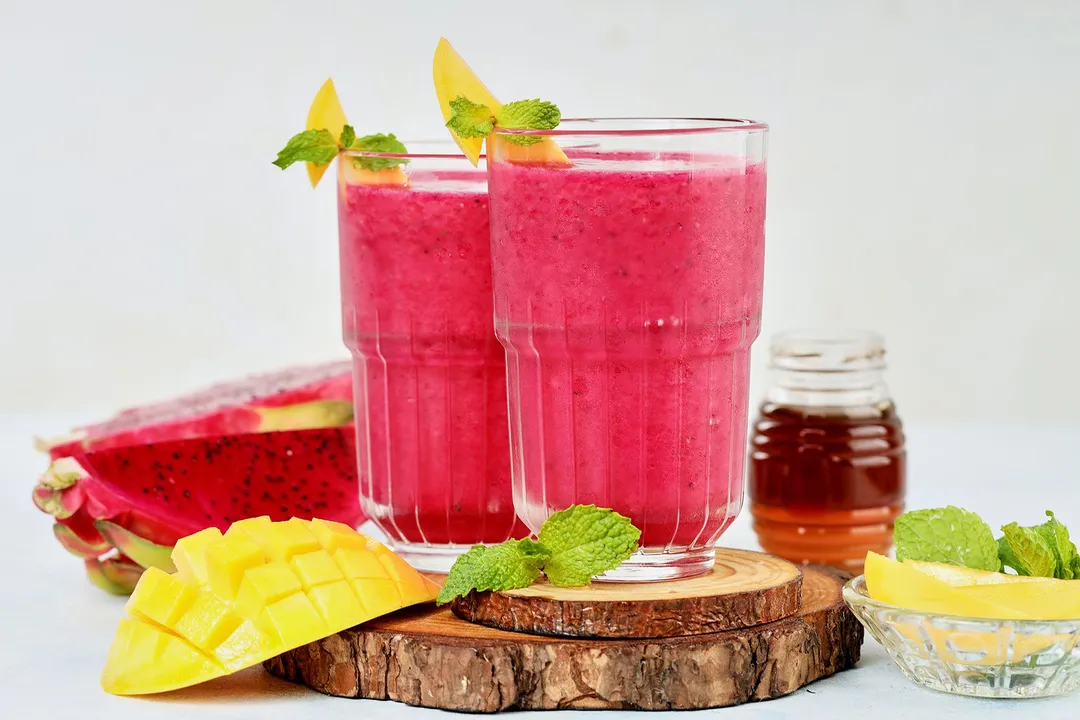 two glasses of mango dragon fruit smoothie garnished with mango sliced, dragon fruit and honey jar