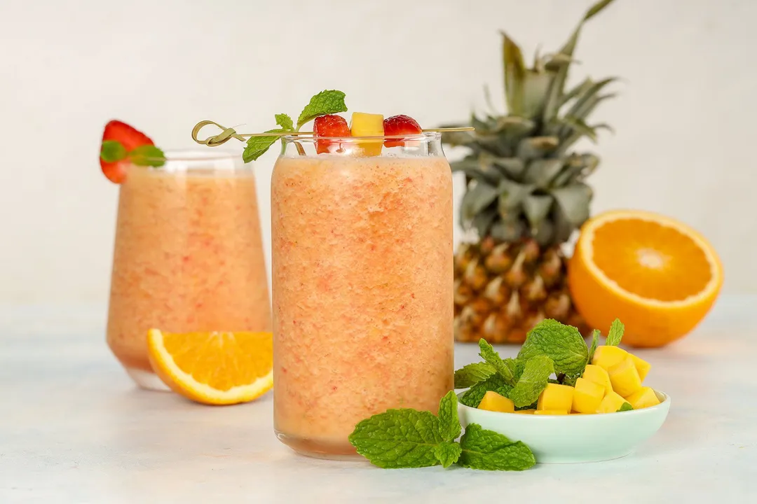 two glasses of smoothie surrounded by pine apple, half orange, mango cubed and mint leaves