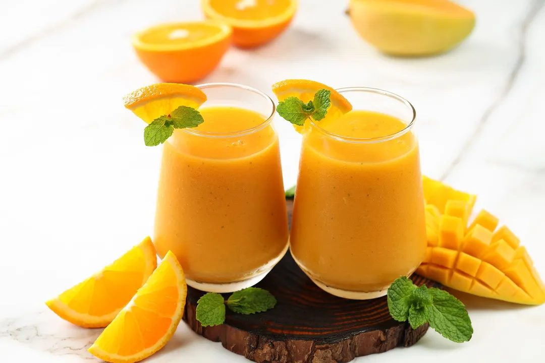 two glasses of yellow smoothie on a tray decorated with mango cubes and orange wedge
