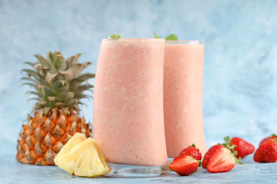 two glasses of strawberry pineapple smoothie surrounded by strawberries, pineapple cubed and pineapple