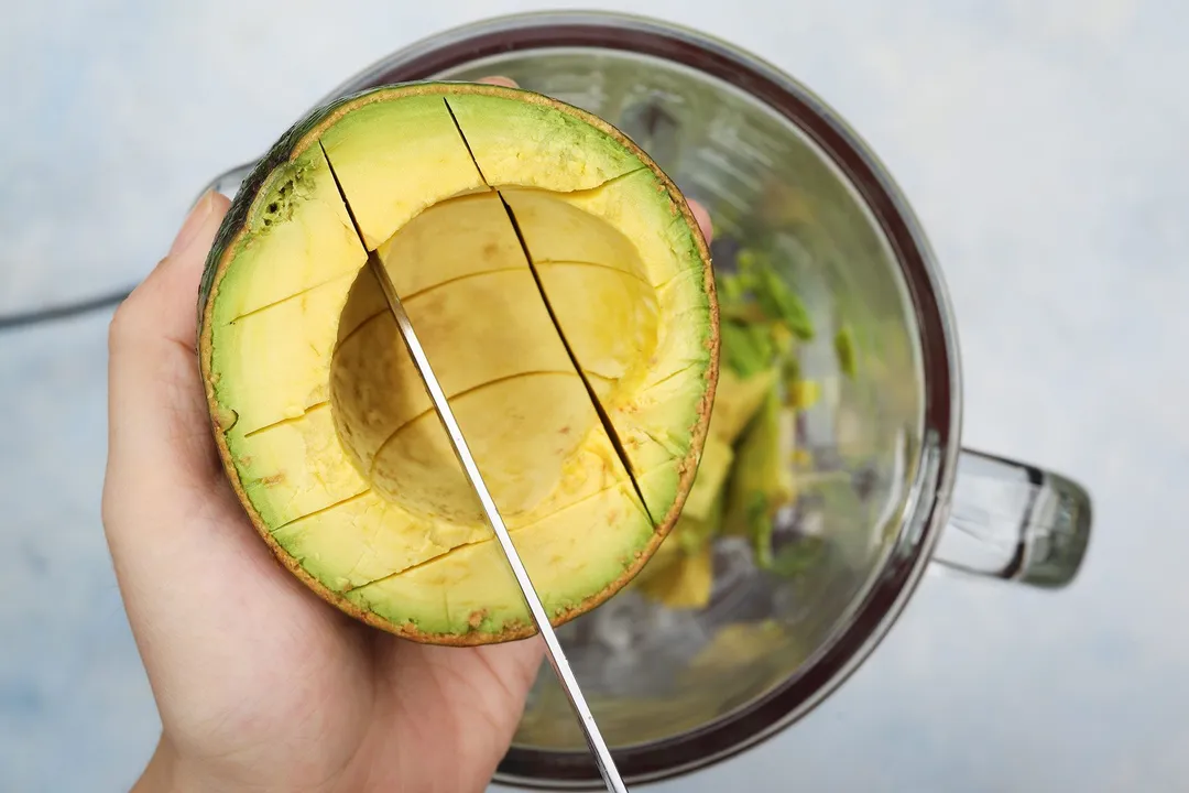 a hand holding a half avocado and a knife cutting into cubes.