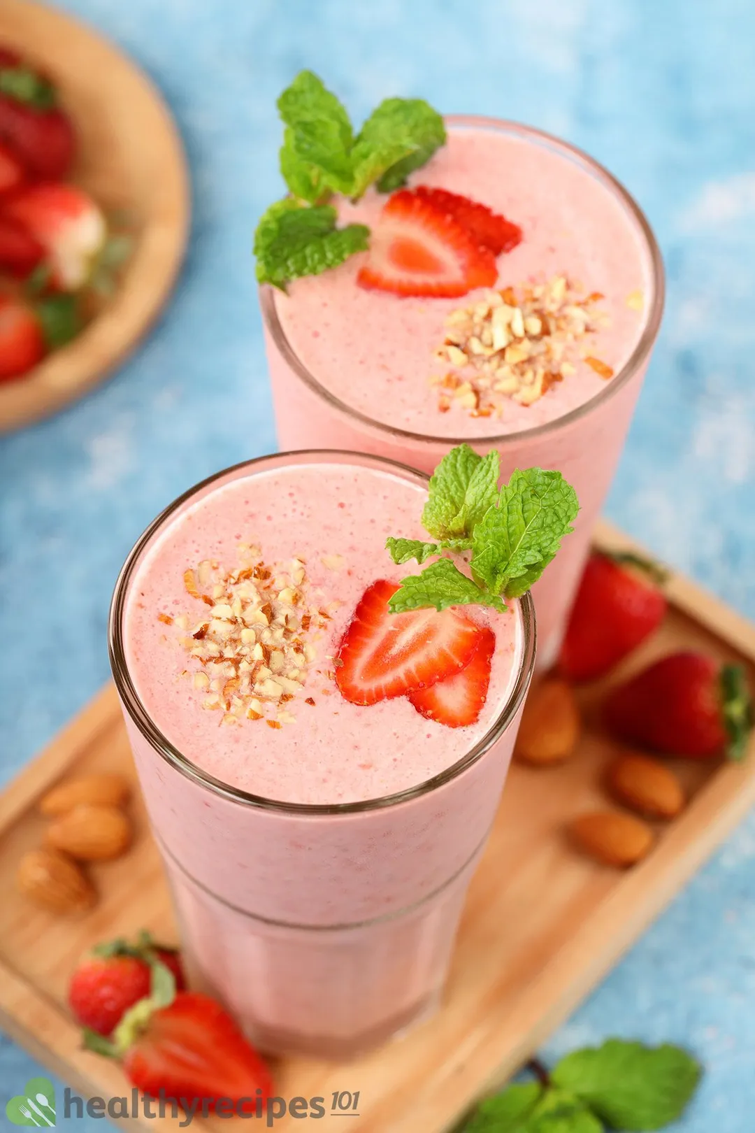 A high angle shot of two glasses of strawberry smoothie laid on a wooden board near strawberry slices and mint leaves.
