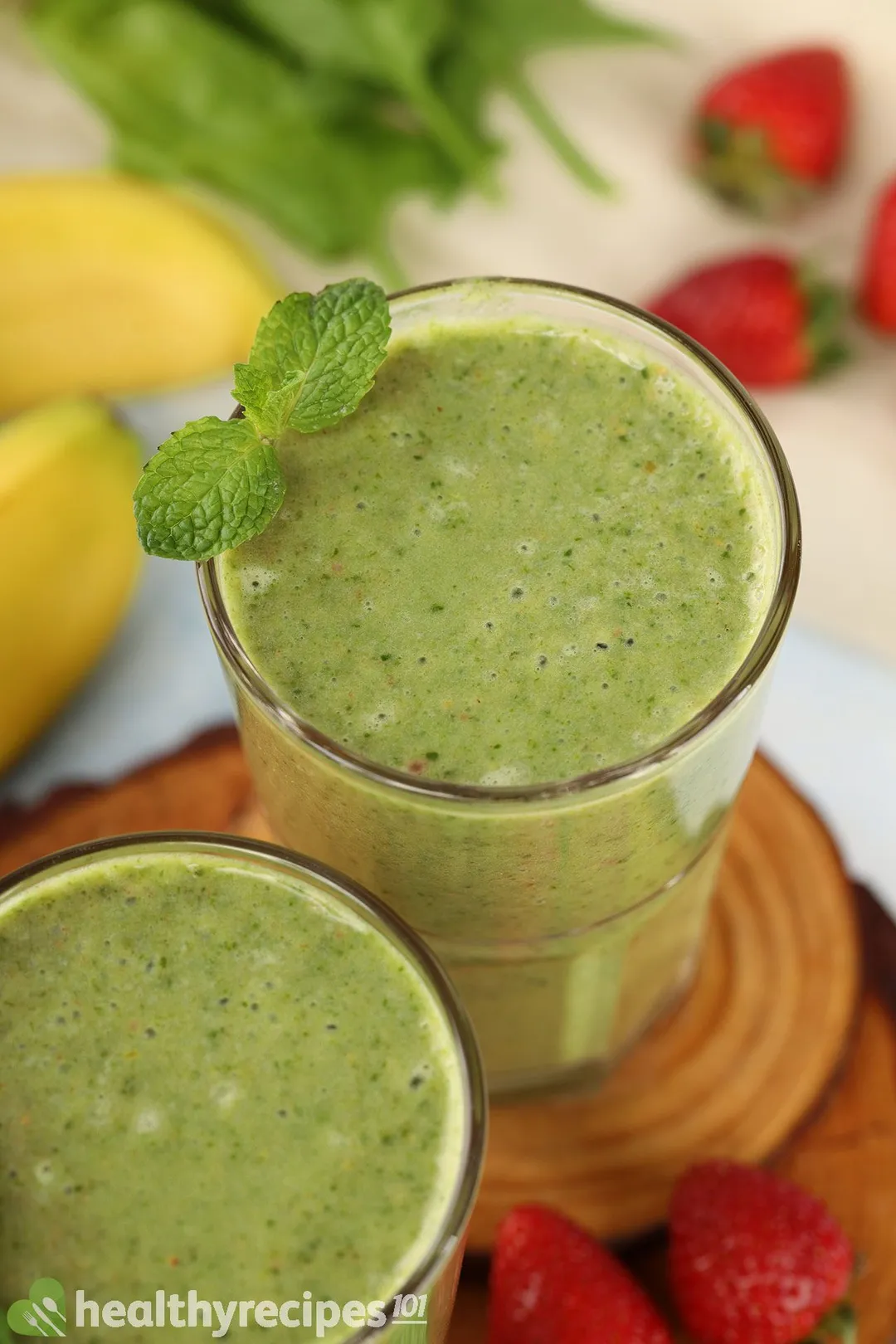 A high-angle shot of two glasses of strawberry spinach banana smoothie laid on a wooden board and near strawberry slices and mint leaves.