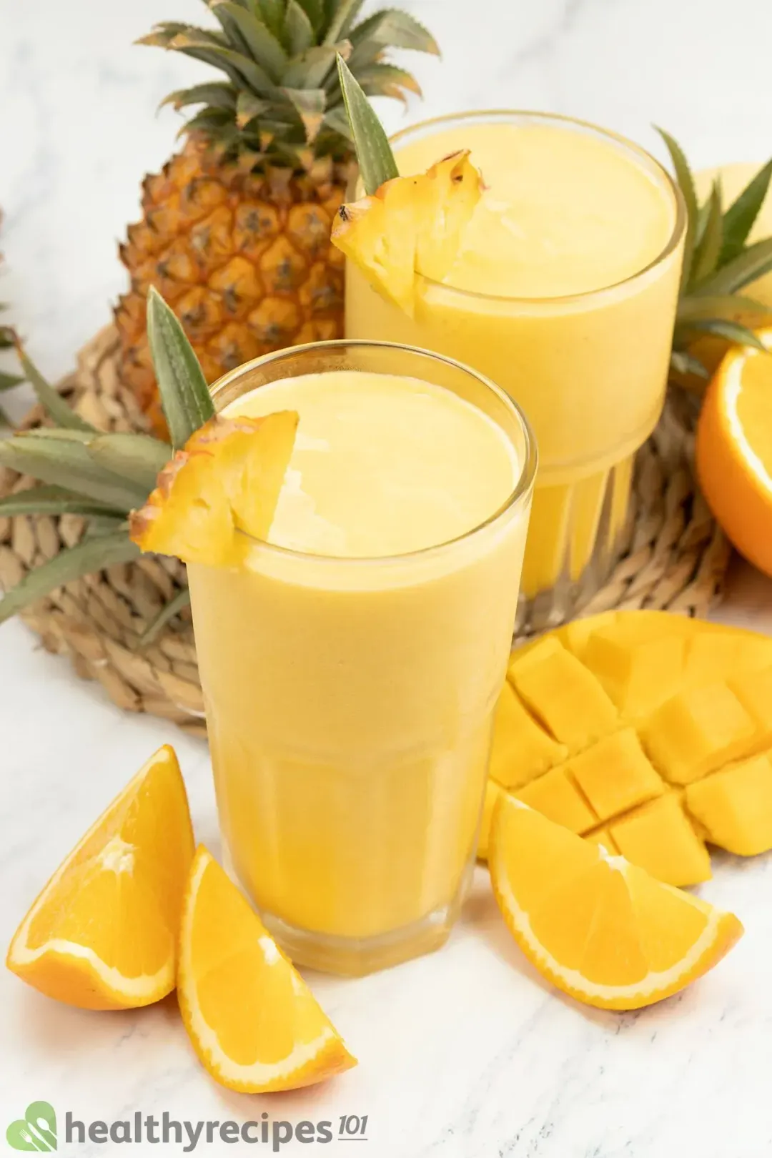 Two glasses of mango smoothie garnished with orange wedges, whole pineapples, and cubed mangoes