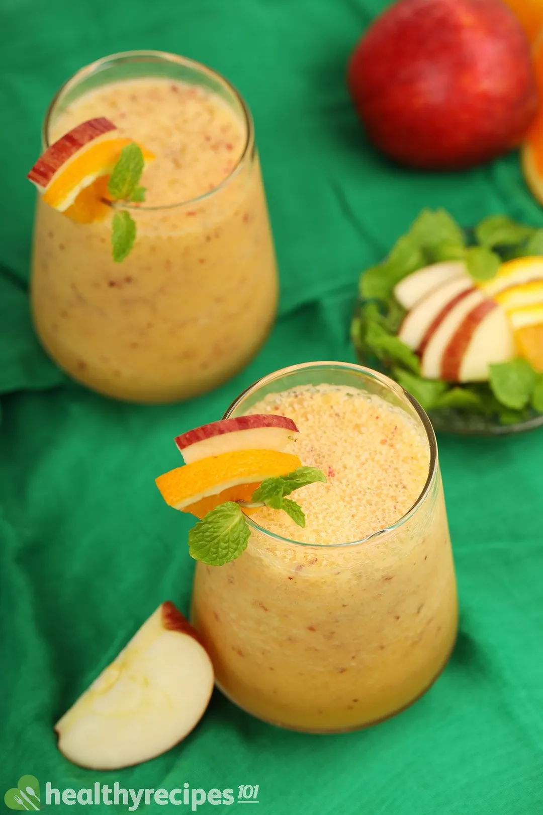 Two glasses of apple orange smoothie and a plate of apple slices laid on a green cloth.