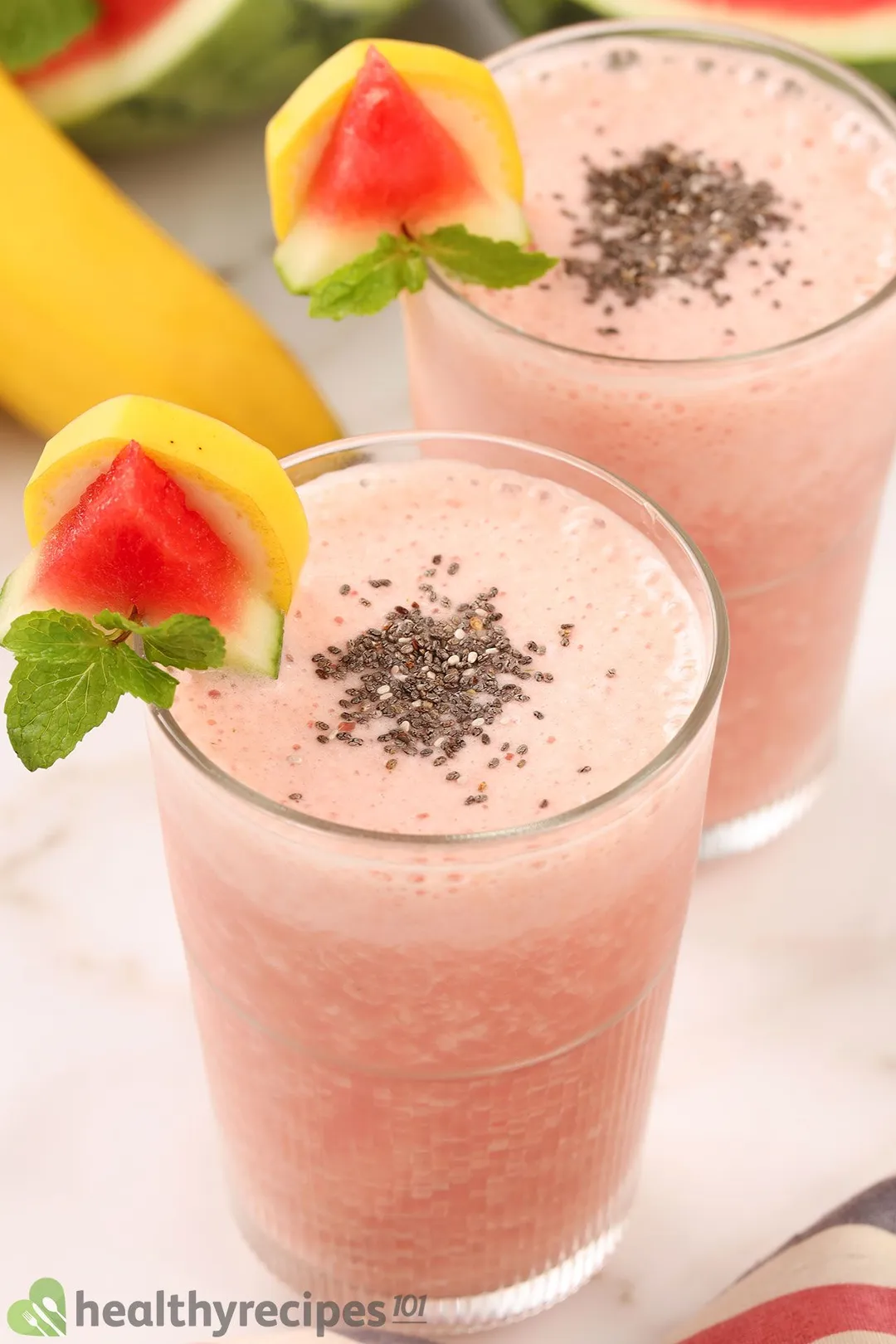 Two glasses of Watermelon Banana Smoothie garnished with chia seeds, mint leaves, a small watermelon wedge, and a piece of banana.