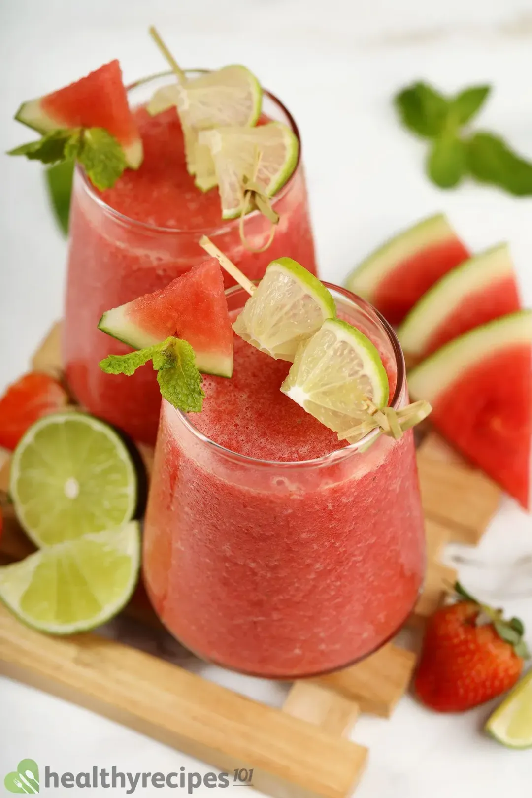 Two glasses of watermelon mojito smoothie decorated with mint leaves, watermelon wedges and lime wedges on a wooden tray