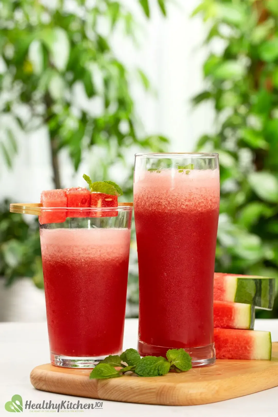 Two glasses of watermelon smoothie decorated with watermelon cubes on a wooden board surrounded by mint leaves and watermelon slices