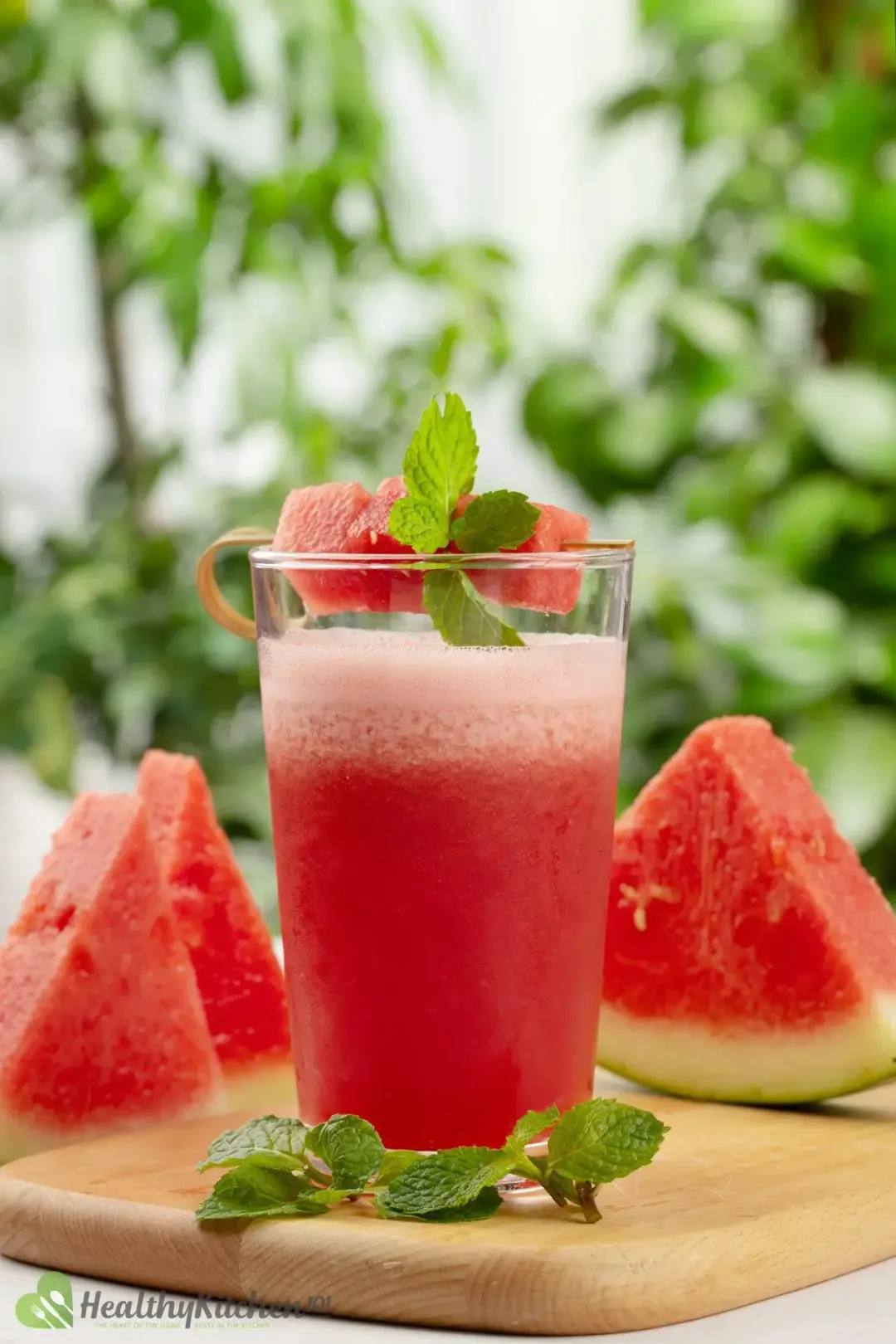 A glass of watermelon smoothie garnished with watermelon cubes and mint leaves on a wooden board next to three watermelon slices and some mint leaves