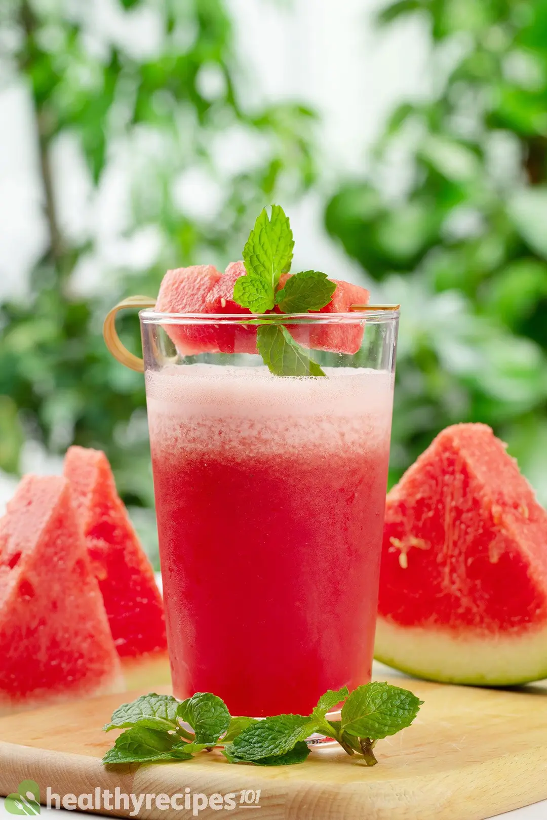 a glass of watermelon smoothie on a wooden tray with watermelon cubed and mint leaves on top, decorated with three slices of watermelon and mint leaves