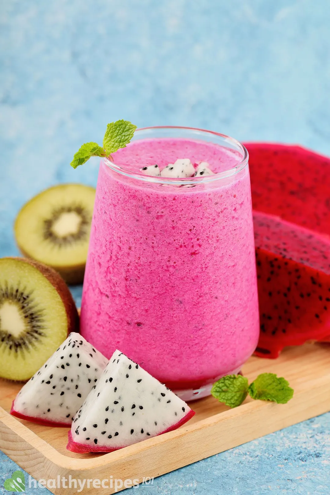 A glass of Dragon Fruit Smoothie placed on a wooden board near dragon fruit triangles, sliced green kiwis, and red dragon fruit.