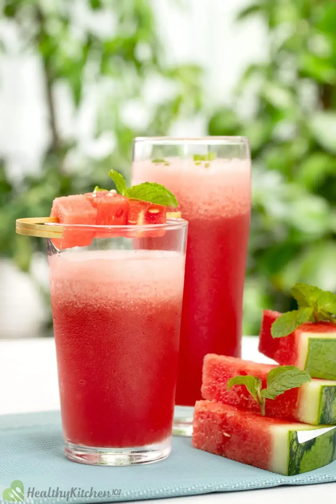 Two glasses of watermelon smoothie with watermelon cubes on top next to three slices of watermelon on a blue cloth