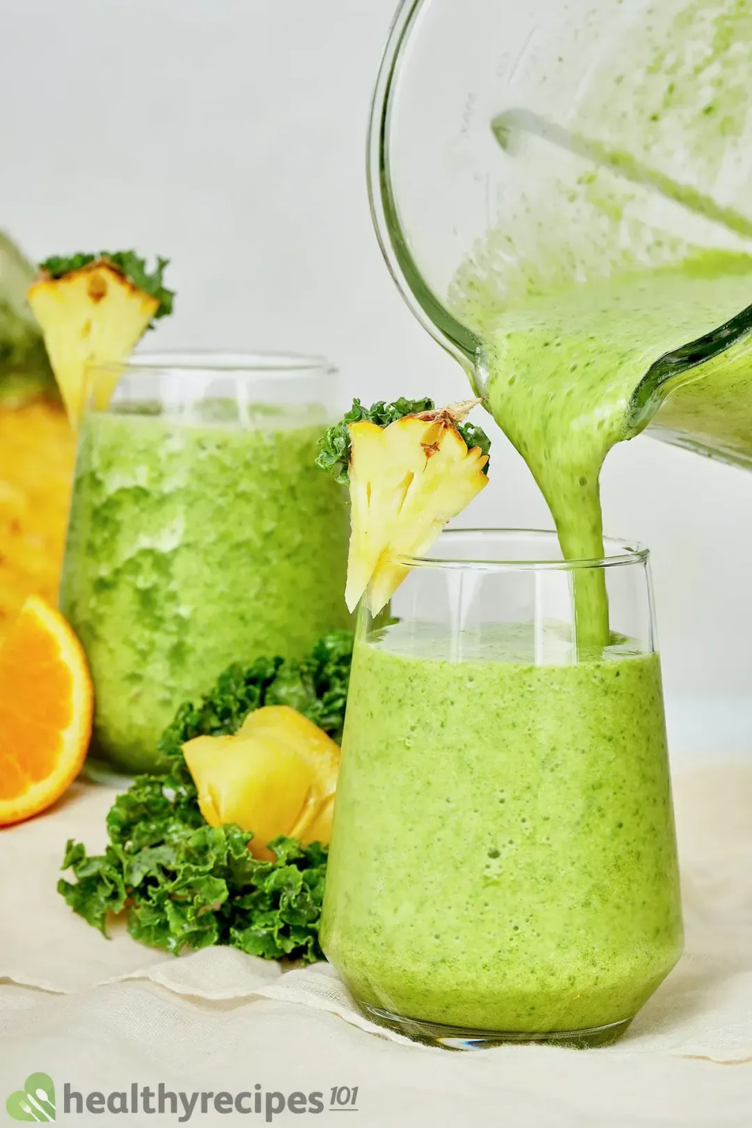 two glasses of pineapple green smoothie, one of which held in the air about to be placed onto a bed of kale and some pineapple wedges