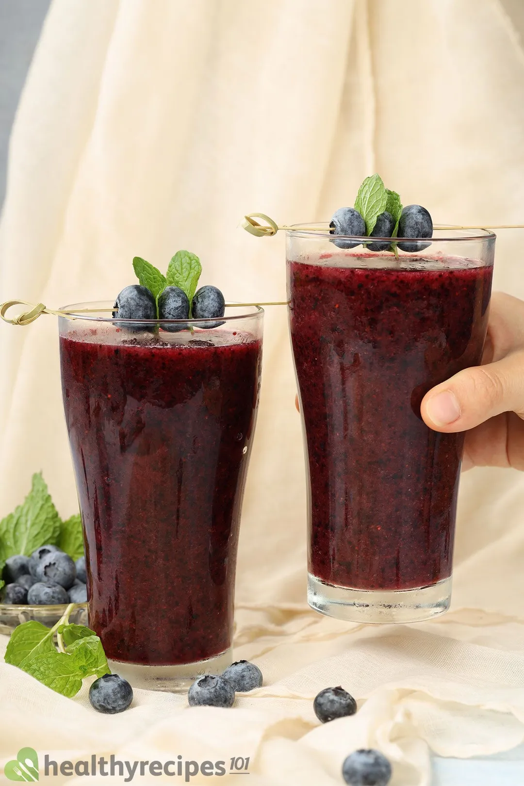 A hand picking up a glass of blueberry smoothie placed next to another identical glass and near some blueberries.