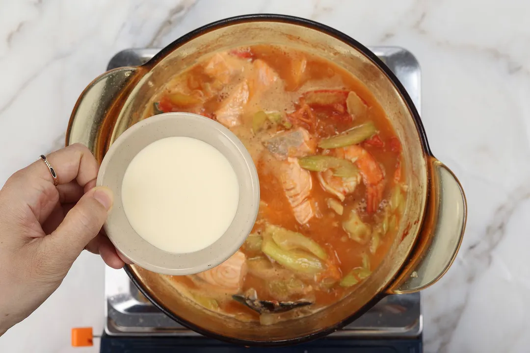 hand holds a small bowl of heavy cream on top of a pot of seafood soup