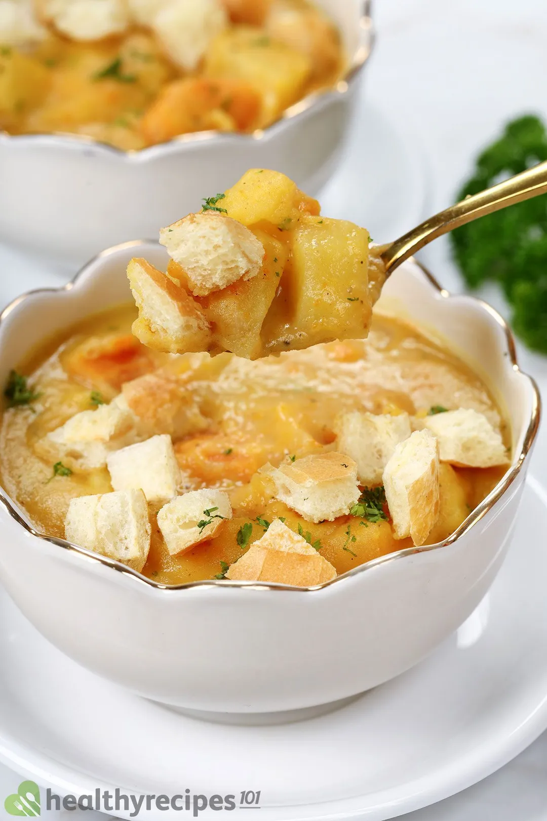 a spoon with potato cubes and croutons on top of a bowl of soup