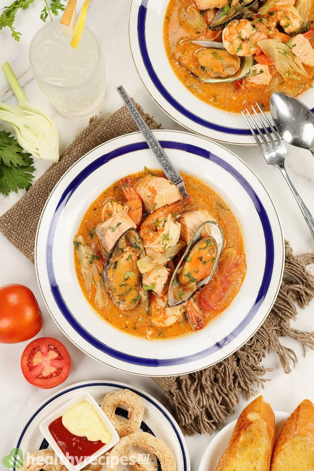 top view of two plate of seafood soup and a plate of onion rings, garlic bread and a glass of juice